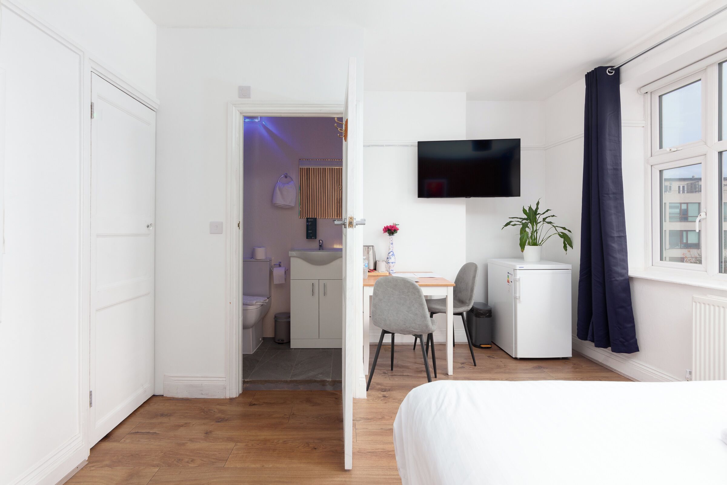 A hotel room with a bed, a desk with two chairs, a small refrigerator, a TV mounted on the wall, and a window with curtains.