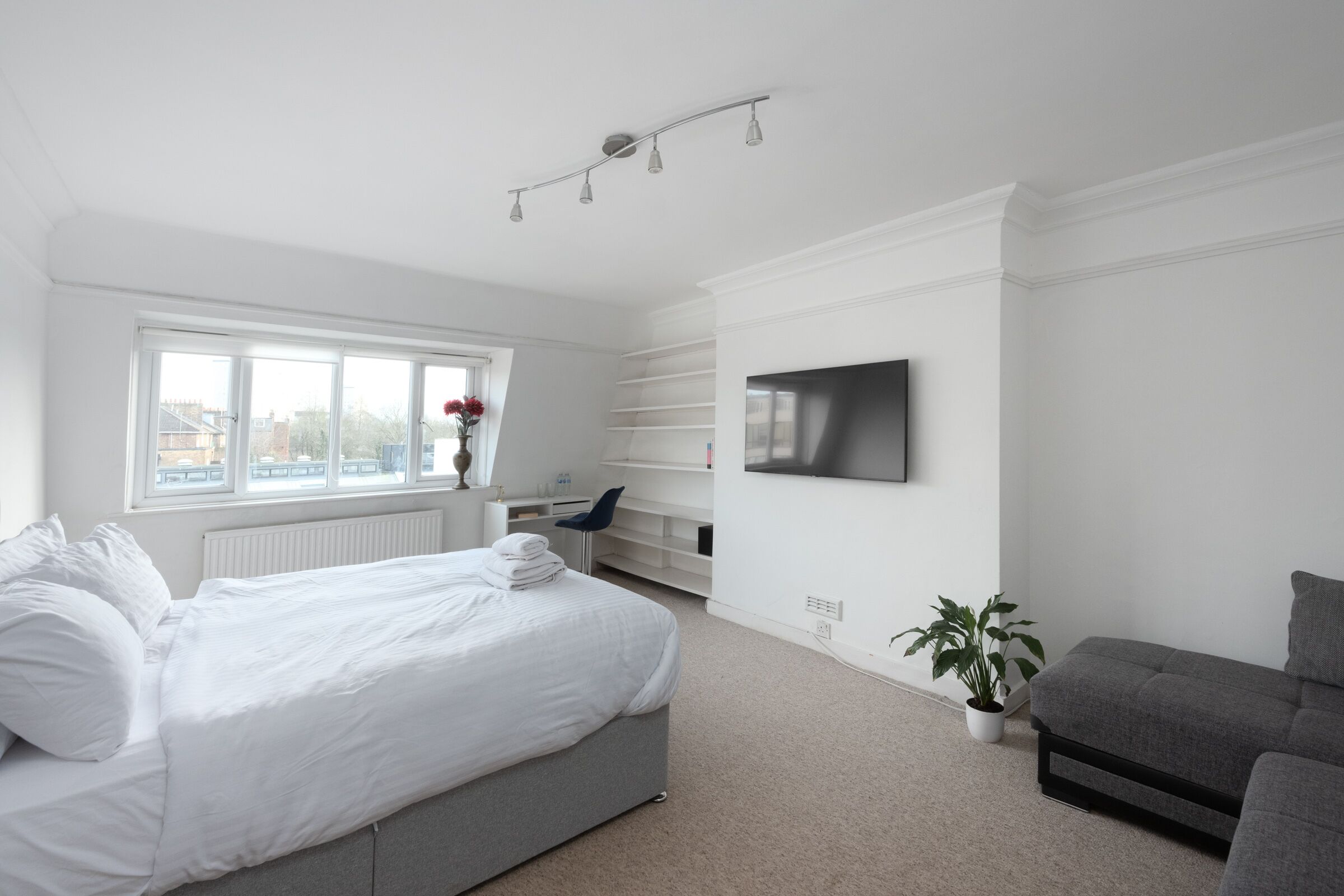 Une chambre moderne avec un grand lit, un canapé gris, un téléviseur fixé au mur et une fenêtre donnant sur des immeubles.