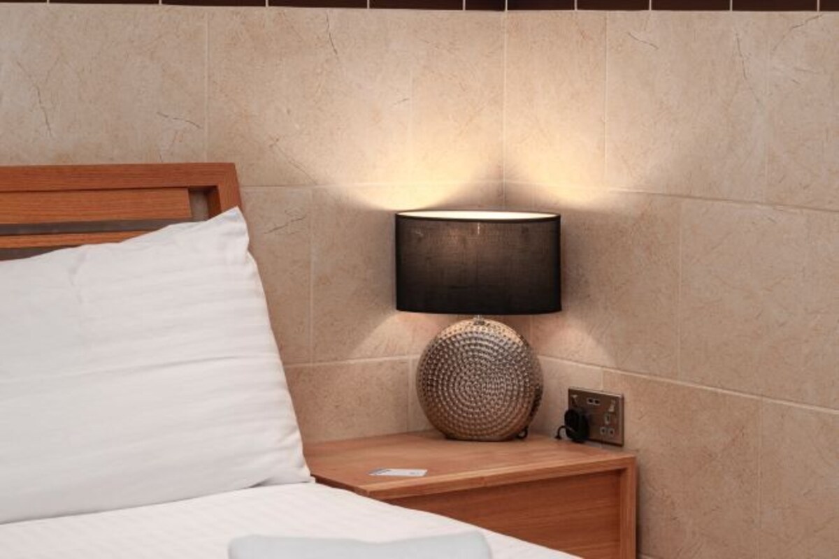 A neatly made bed with white linens, a wooden headboard, a bedside lamp with a textured lampshade, and a small wooden nightstand with a power outlet.