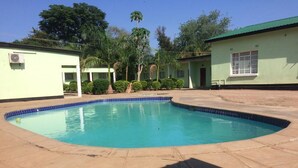 A swimming pool with a curved edge, surrounded by a paved area and a building with a green roof. - Richland Lodge Motel (Livingstone)