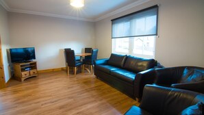A living room with a wooden floor, a black leather sofa, a TV on a wooden stand, and a window with blinds. - Morris Gardens Apartments (Inverurie)