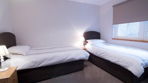 A room with two single beds, a bedside table with a lamp, a window with a blind, and a carpeted floor. - Morris Gardens Apartments (Inverurie)