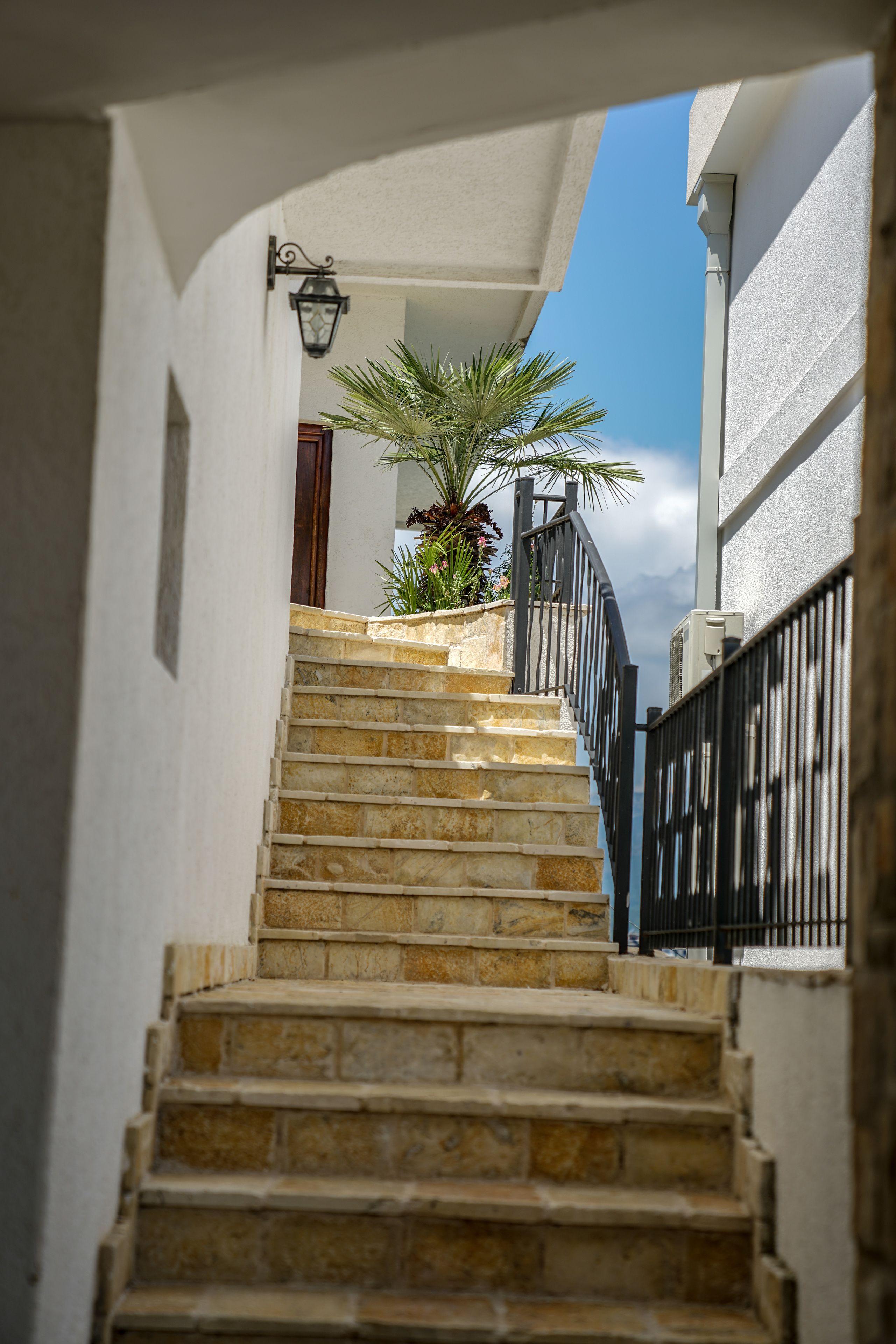 Un escalier en pierre mène à un bâtiment blanc, orné d’une balustrade noire et surmonté d’un pot de fleurs.
