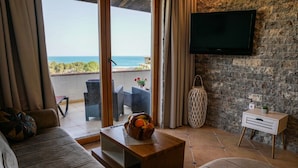 A living room with a sofa, a coffee table with fruit, a TV on a stone wall, and a view of the sea. - Apartments Bazar (Ulcinj)