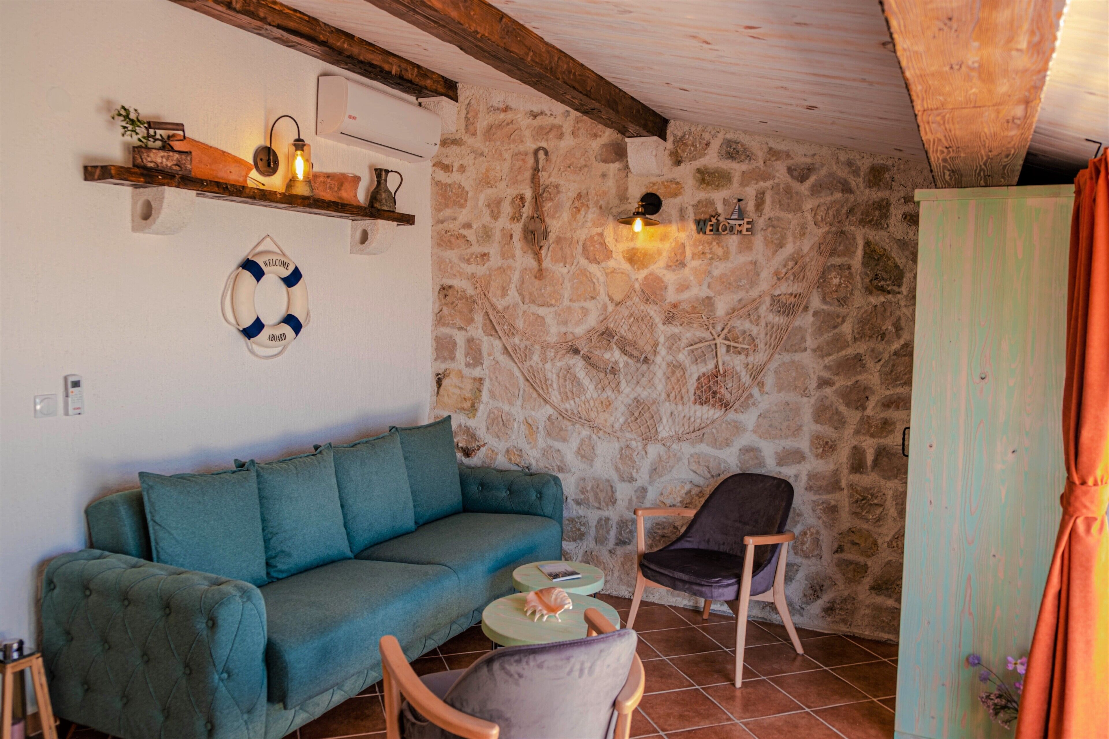 A cozy room with a stone wall, a teal sofa, a wooden chair, and a small round table with a book and a shell.