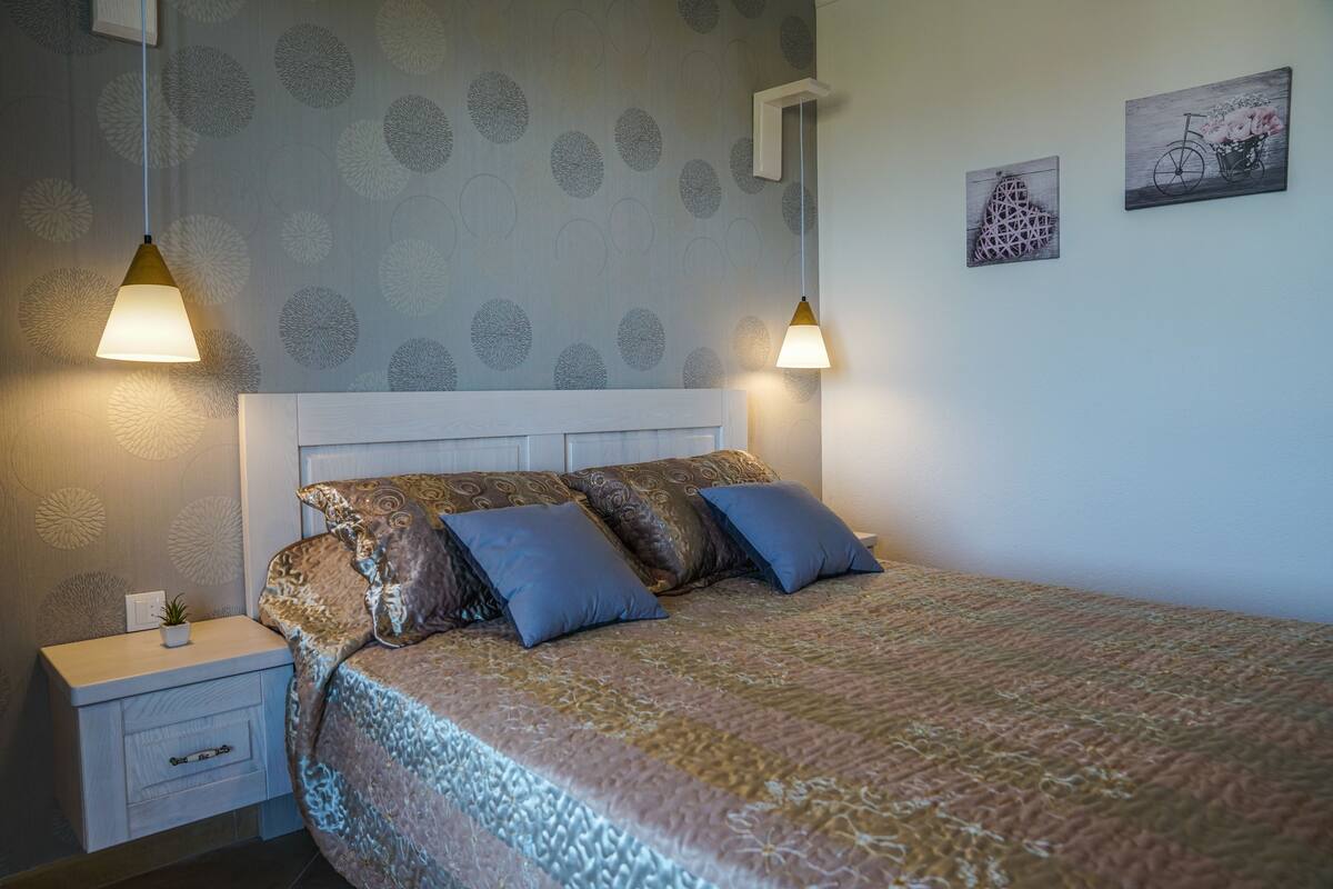 Ein Schlafzimmer mit einem gemusterten Bettbezug, zwei Nachttischlampen, einem Nachttisch mit einer Pflanze und zwei an der Wand befestigten Kunstwerken.