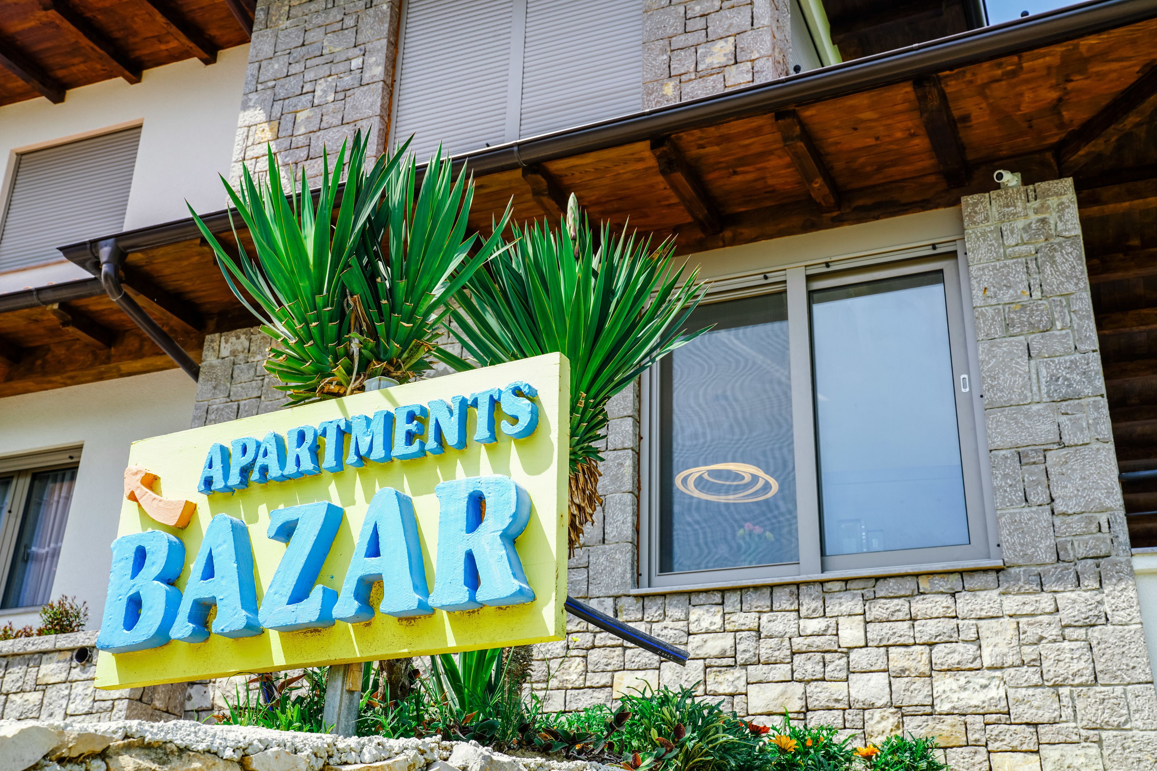 Señal para 'Apartments Bazaar', con un edificio de piedra y un techo de madera al fondo.