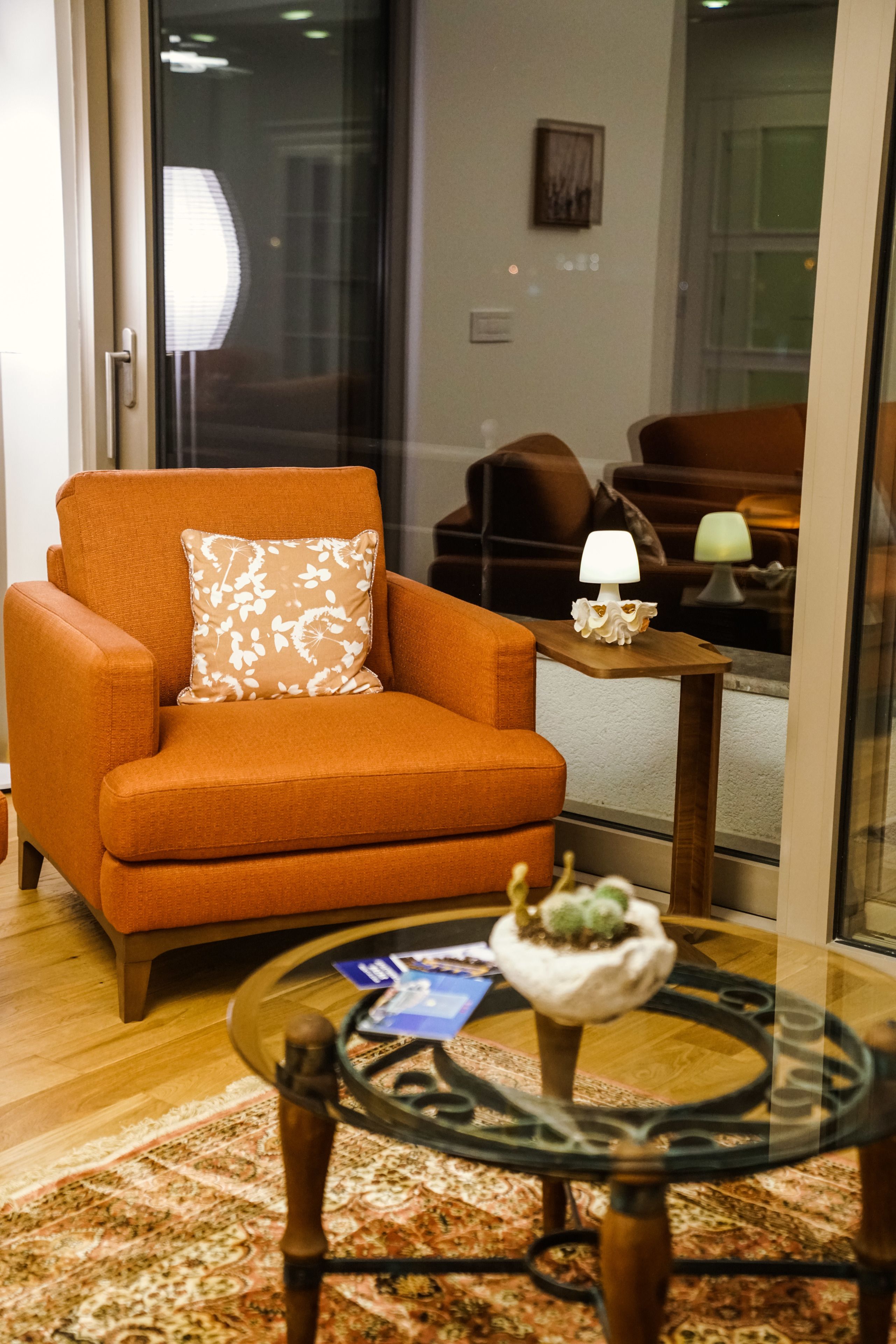 A modern living room with an orange armchair, a glass coffee table with a decorative item, and a small wooden side table with a lamp.