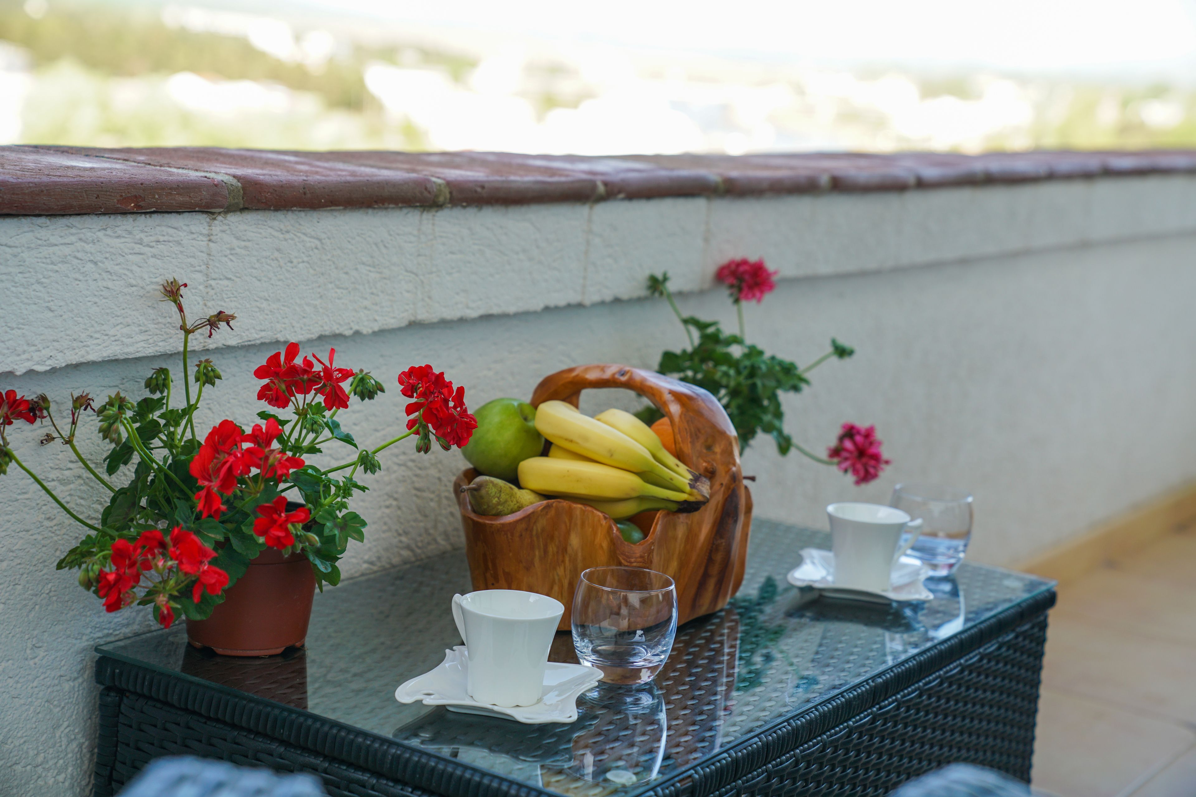 A table with a potted plant, a wooden bowl of fruit, and two cups on a glass table.