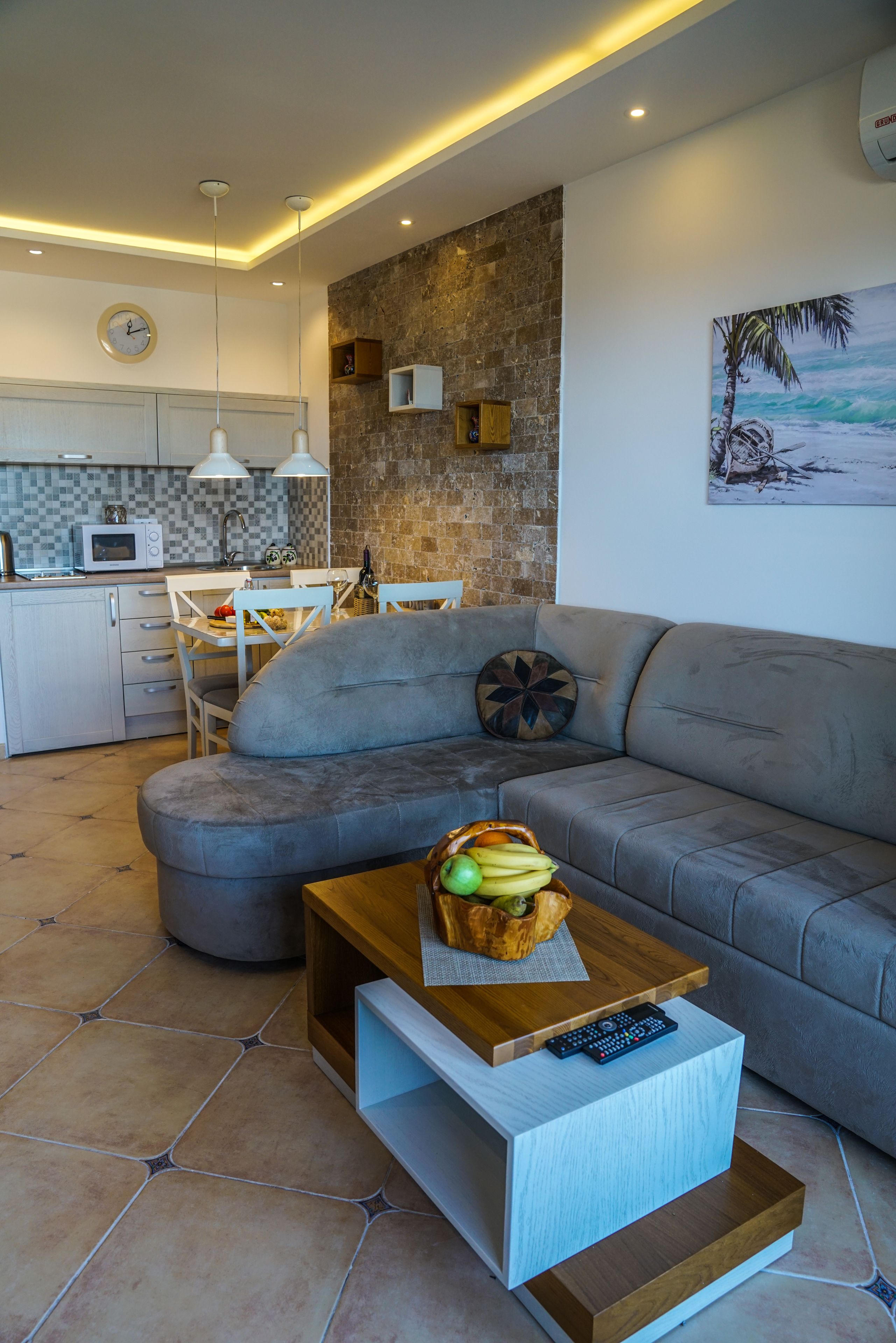 A modern living room with a grey sofa, a wooden coffee table with a fruit bowl, a remote control, and a kitchen in the background.