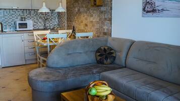 A modern living room with a grey sofa, a wooden coffee table with a fruit bowl, a remote control, and a kitchen in the background.