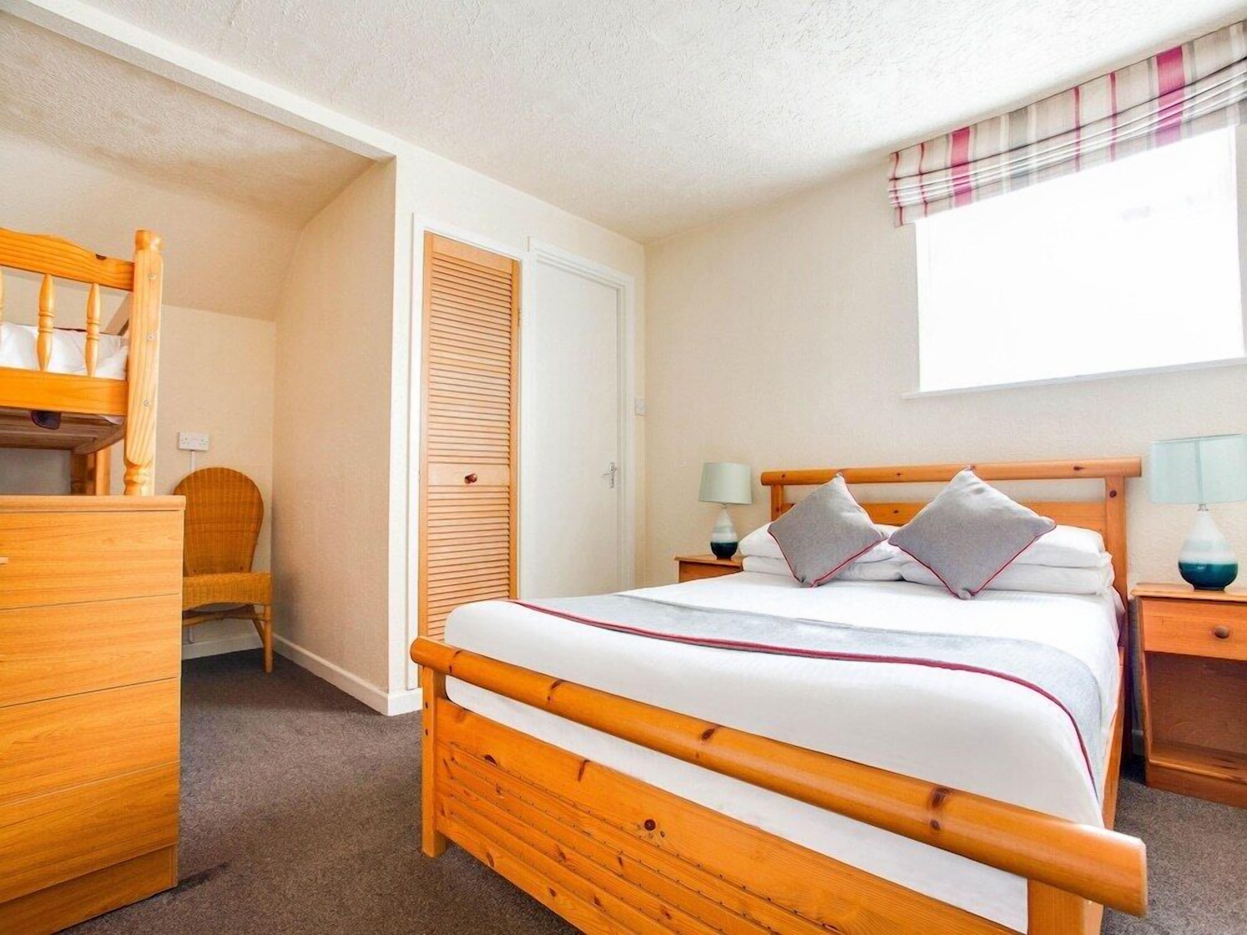 Een slaapkamer met een houten bed, een dressoir, een stoel, een nachtkastje met een lamp, en een raam met een geruit gordijn.