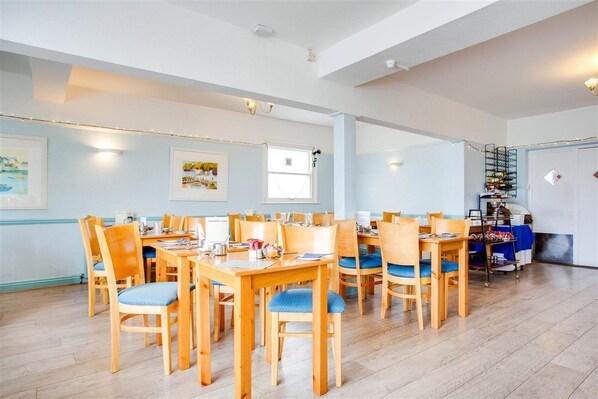 A spacious dining area with multiple wooden tables and chairs, a staircase with a railing, and a wall-mounted light fixture. - Godolphin Arms Hotel (Newquay)