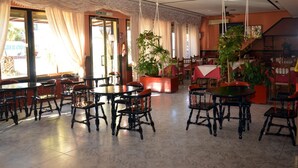 Un restaurante con mesas y sillas redondas, ventanas amplias con cortinas y plantas en macetas.