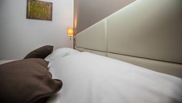 Un lit avec une literie blanche et un oreiller marron, une lampe fixée au mur et un petit tableau accroché au mur.
