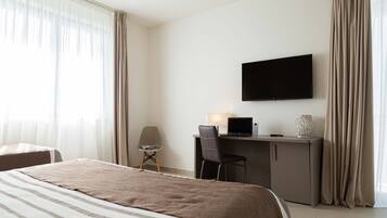 A hotel room with a bed, a desk with a laptop, a chair, a TV on the wall, and curtains.
