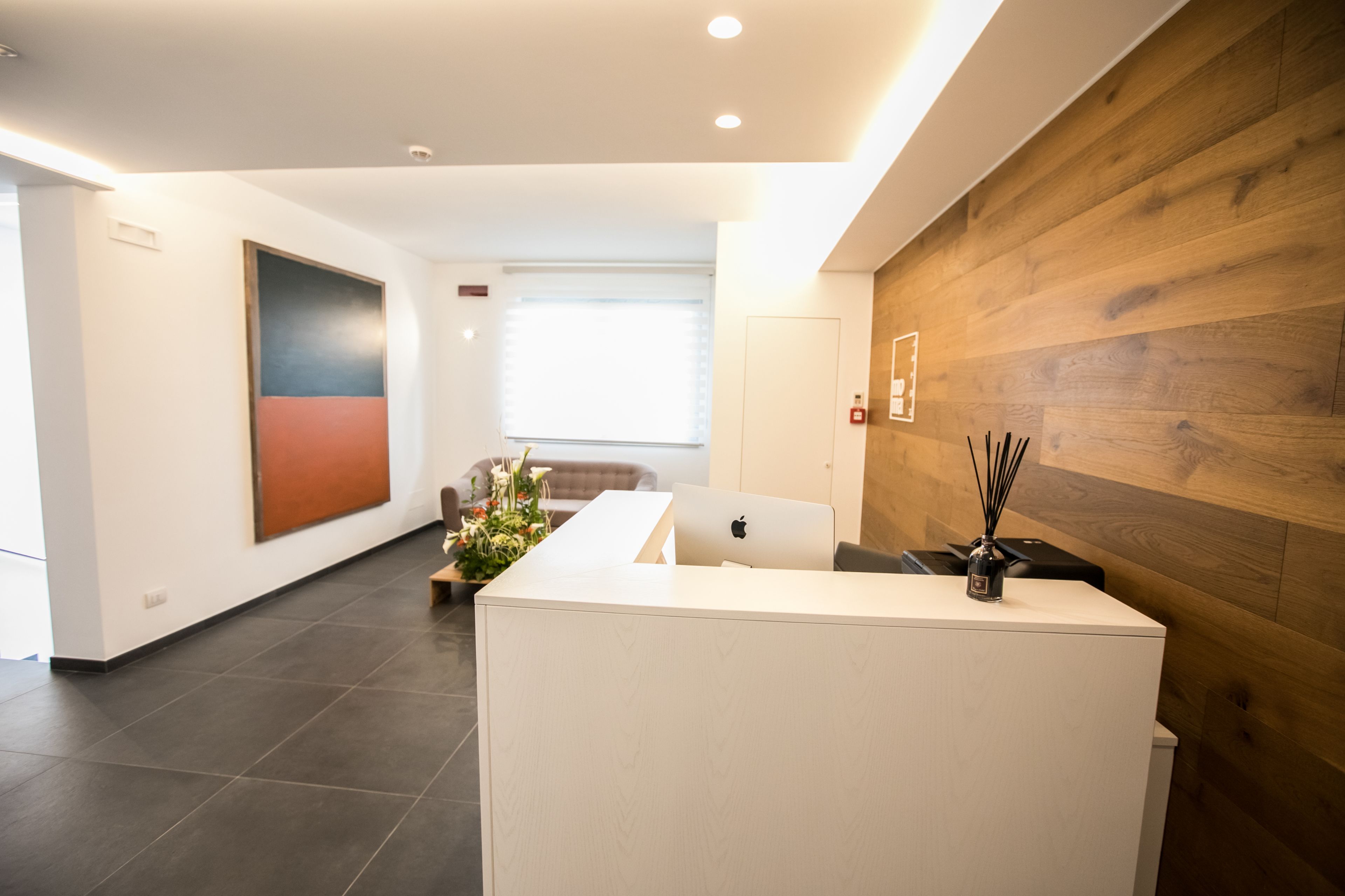 A reception area with a white desk, a laptop, and a vase with reeds. There is a large abstract painting on the wall and a window with blinds in the background.