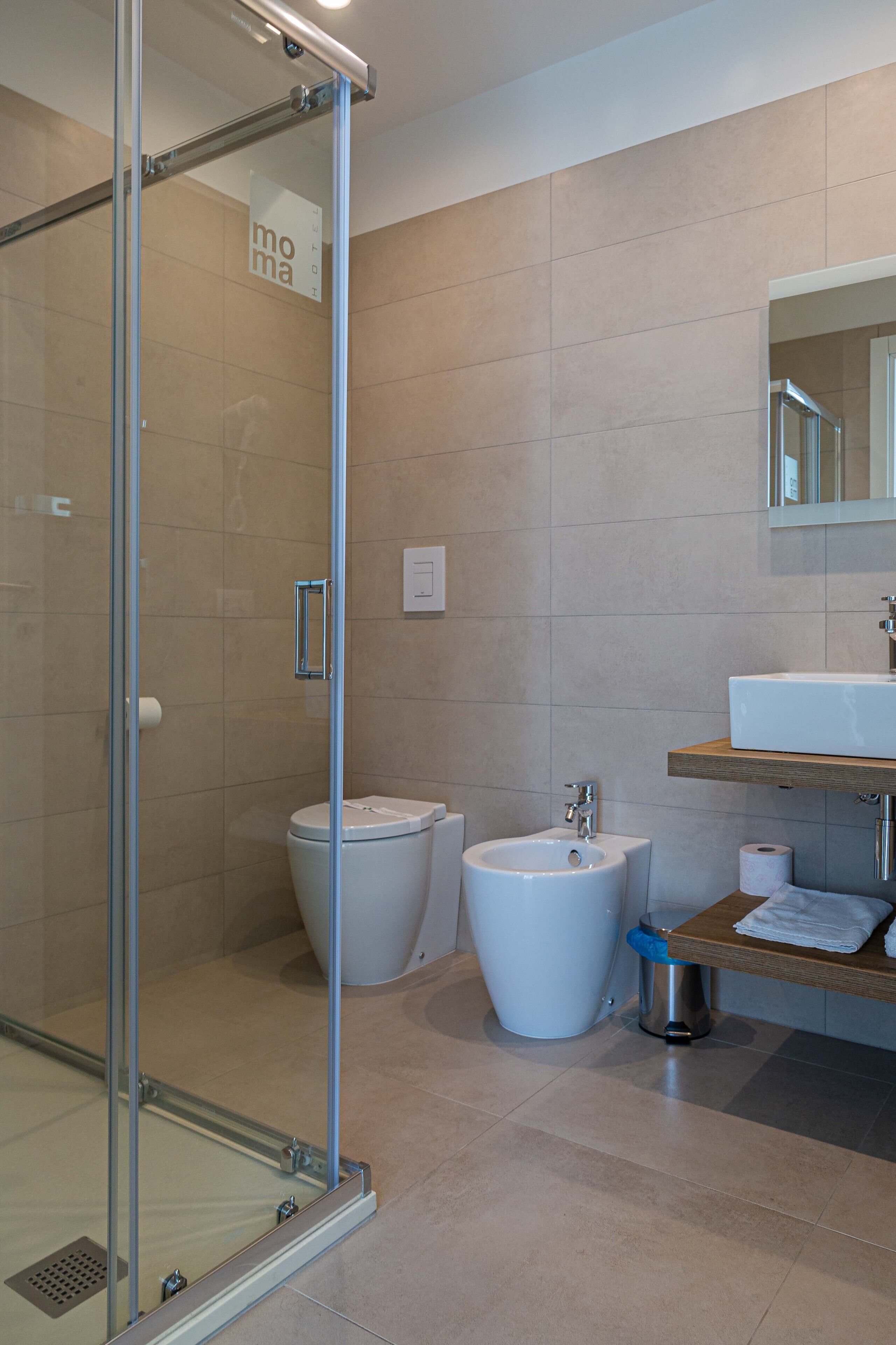 A modern bathroom with a glass shower enclosure, a white toilet, a bidet, and a sink with a wooden countertop.
