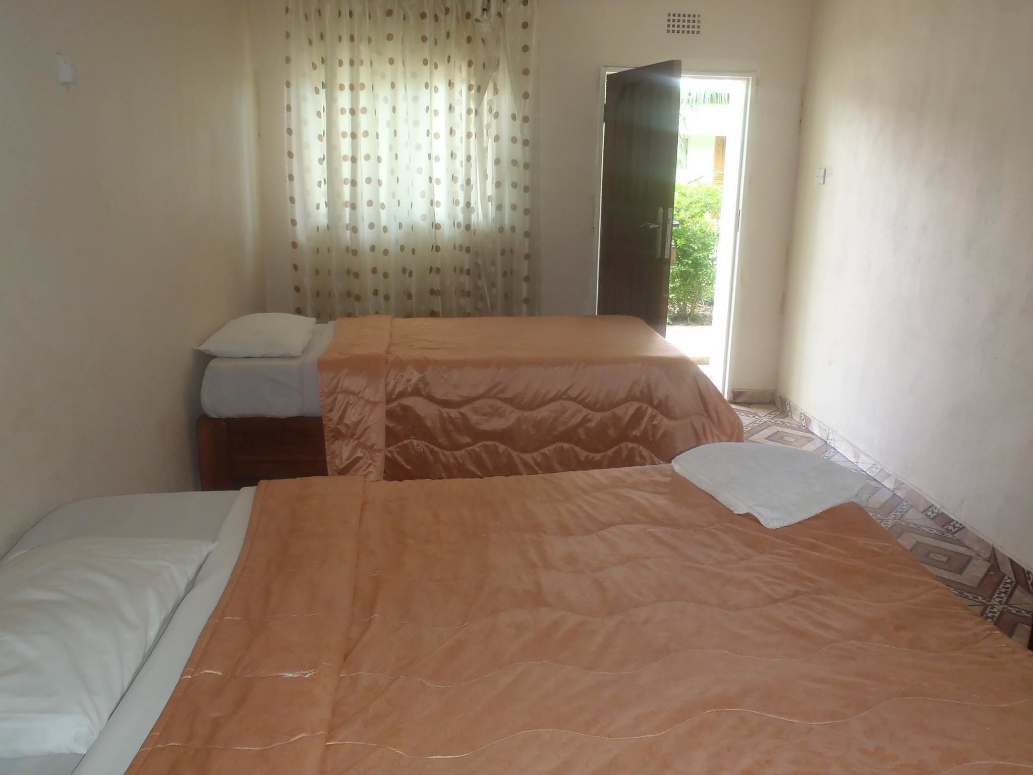 Una habitación con dos camas, una con colcha naranja y la otra con sábanas blancas. Hay una ventana con cortinas y una puerta que da al exterior.