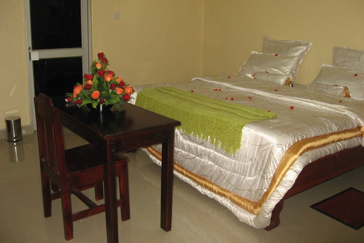 A hotel room with a bed, a green throw blanket, and a vase of flowers on a table.