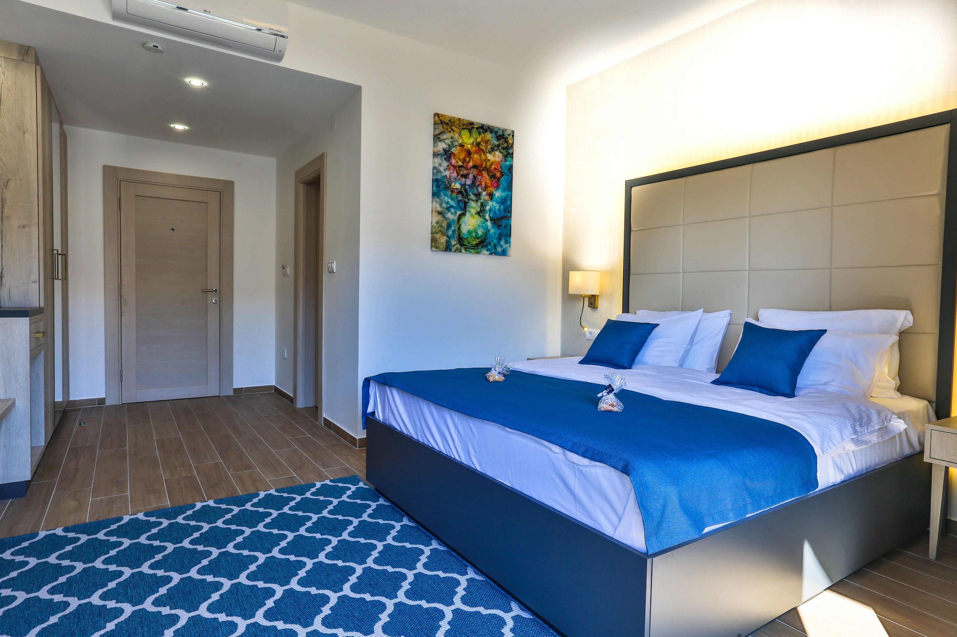 A modern hotel room with a large bed, a blue patterned rug, a painting on the wall, and a wooden wardrobe.