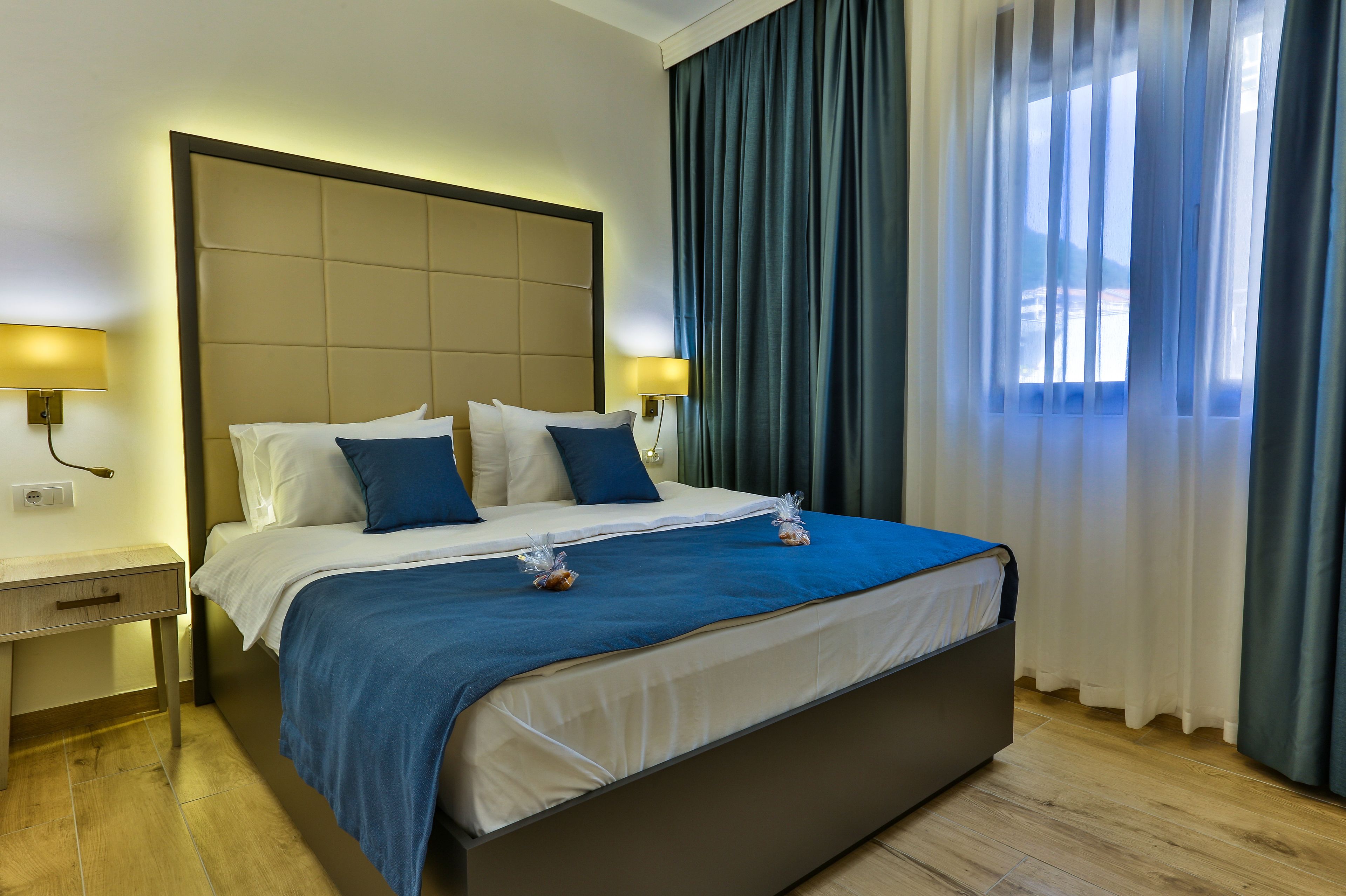 Habitación de hotel con una cama grande, dos mesitas de noche con lámparas y vistas al exterior a través de cortinas translúcidas.