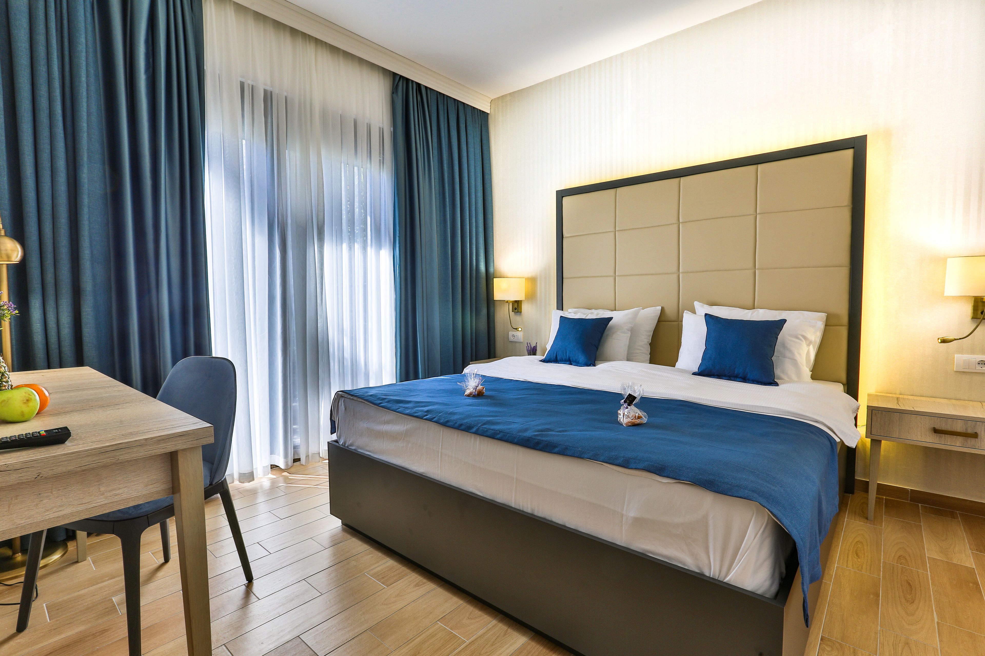 A hotel room with a large bed, two bedside tables with lamps, a desk with a chair, and a window with curtains.