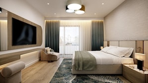 A modern hotel room with a large flat-screen TV mounted on the wall, a comfortable bed with a green throw blanket, a seating area with a sofa and armchair, and a large window with curtains. - Hotel Harmony by Aycon (Petrovac)