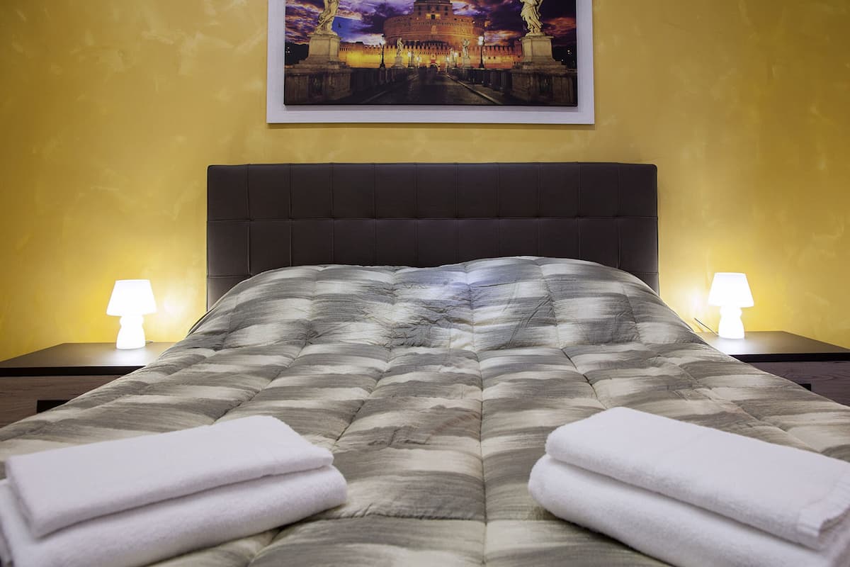 Ein ordentlich bezogenes Bett mit einem gemusterten Überwurf, zwei Nachttischlampen und einem gerahmten Bild darüber.