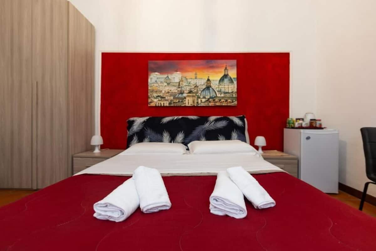 Ein Hotelzimmer mit einem großen Bett, roten Wänden, einem Stadtbildgemälde, zwei Nachttischen mit Lampen und einem kleinen Kühlschrank.