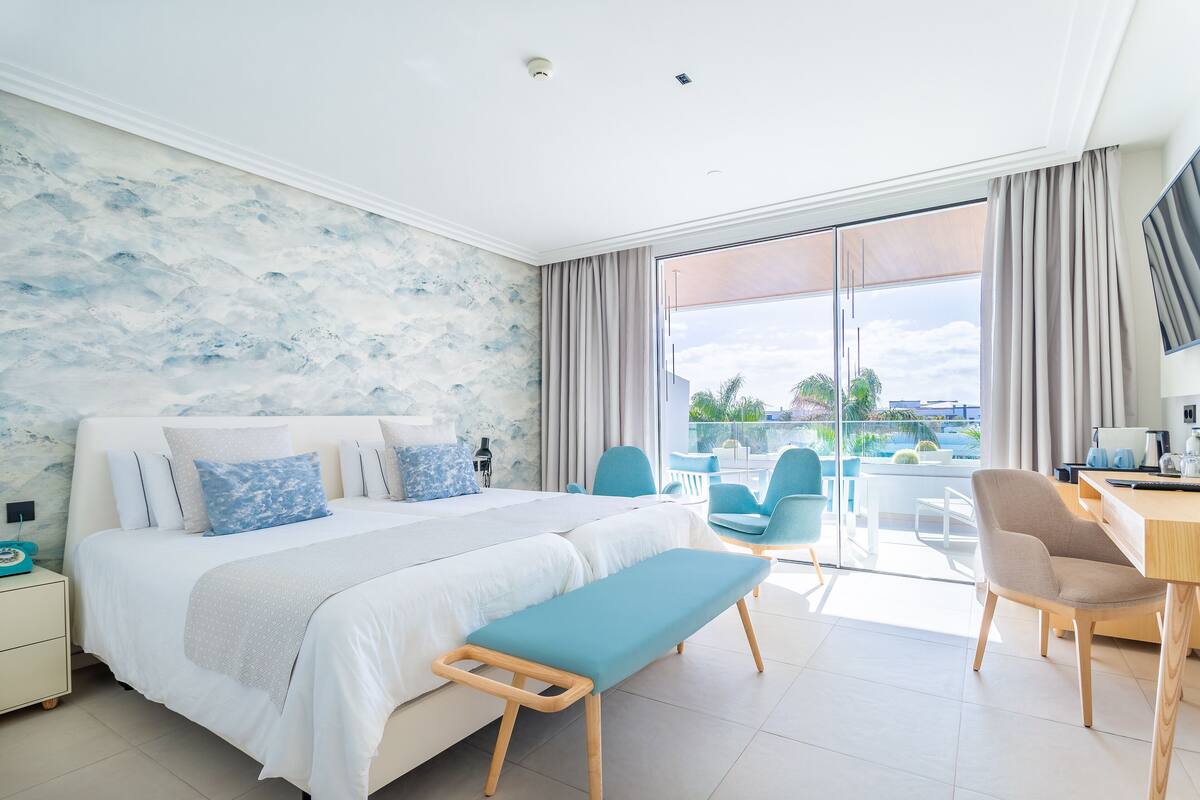 Une chambre d’hôtel moderne avec un grand lit, une banquette, un bureau et un balcon équipé de chaises.