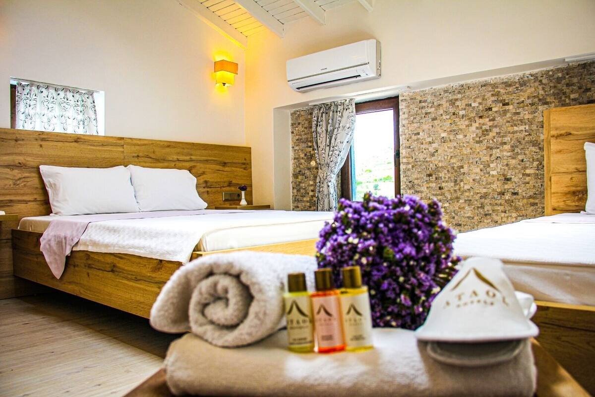 A hotel room with a wooden bed, a small bedside table with toiletries, a window with curtains, and a brick-patterned wall.