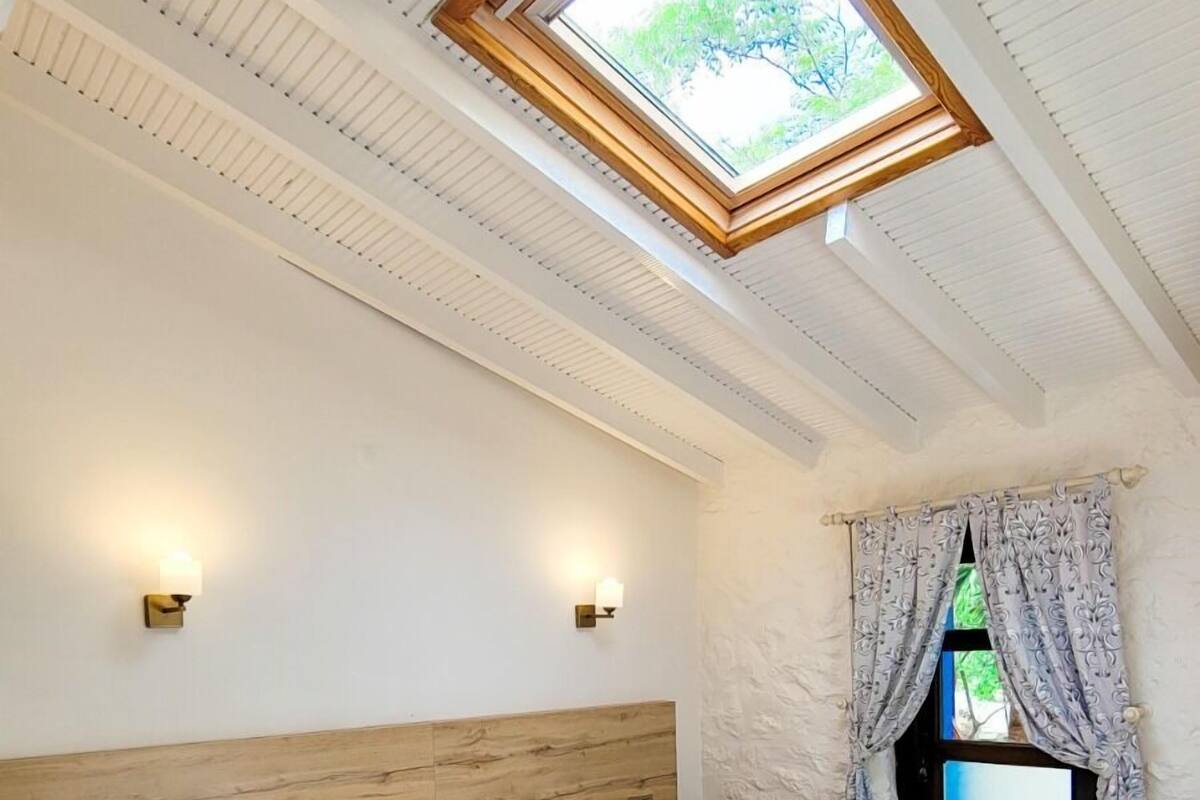 A bedroom with a skylight, wooden headboard, bedside lamps, and a view of greenery outside.