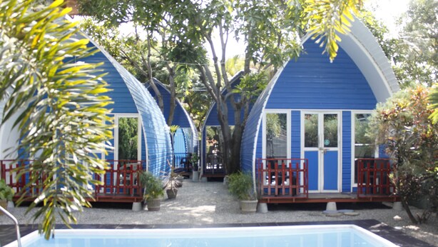 Filas de cabañas azules con barandillas rojas, una piscina y vegetación circundante.