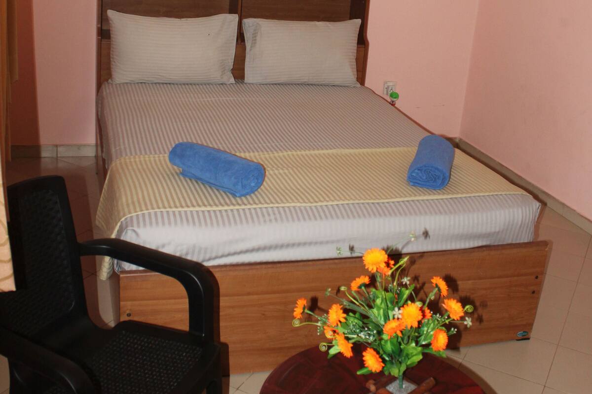 A bedroom with a bed, a chair, a small table with a vase of flowers, and a wall-mounted light.