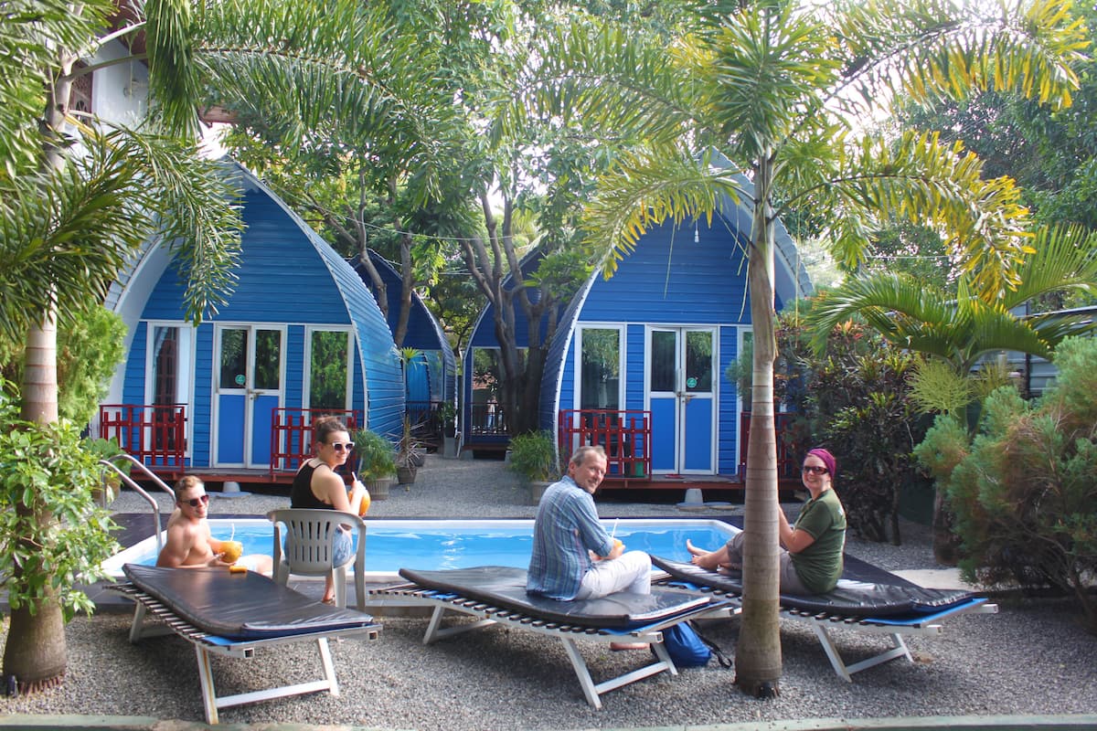 A poolside area with lounge chairs and a swimming pool, surrounded by blue buildings and tropical plants.