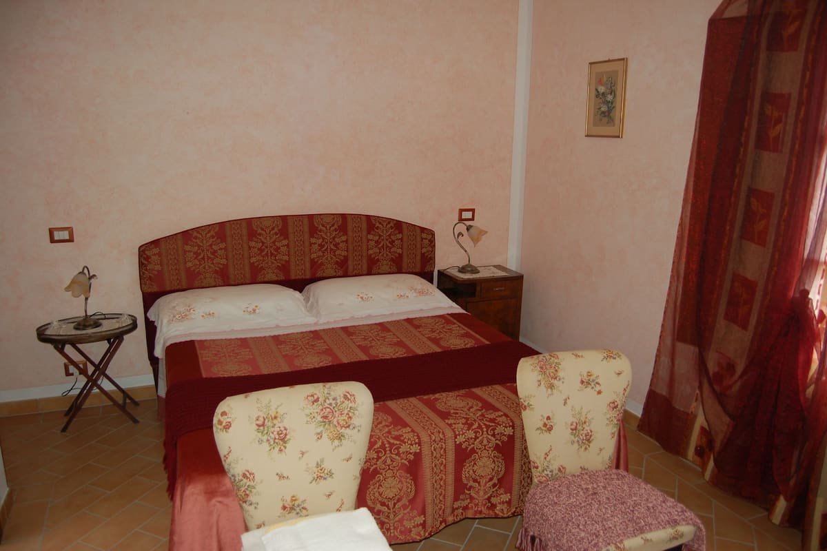 A bedroom with a bed, two chairs, a nightstand, a lamp, and a curtain.