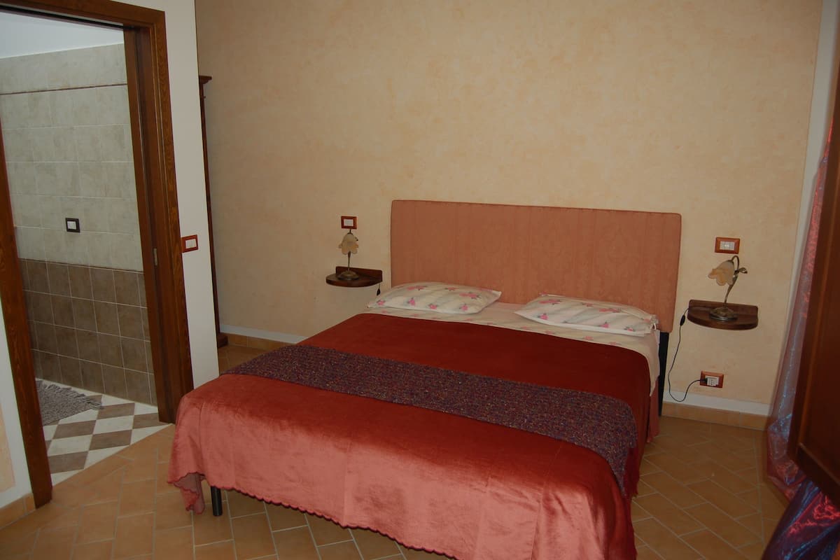 A bedroom with a large bed, two bedside tables with lamps, a door to a bathroom, and a tiled floor.