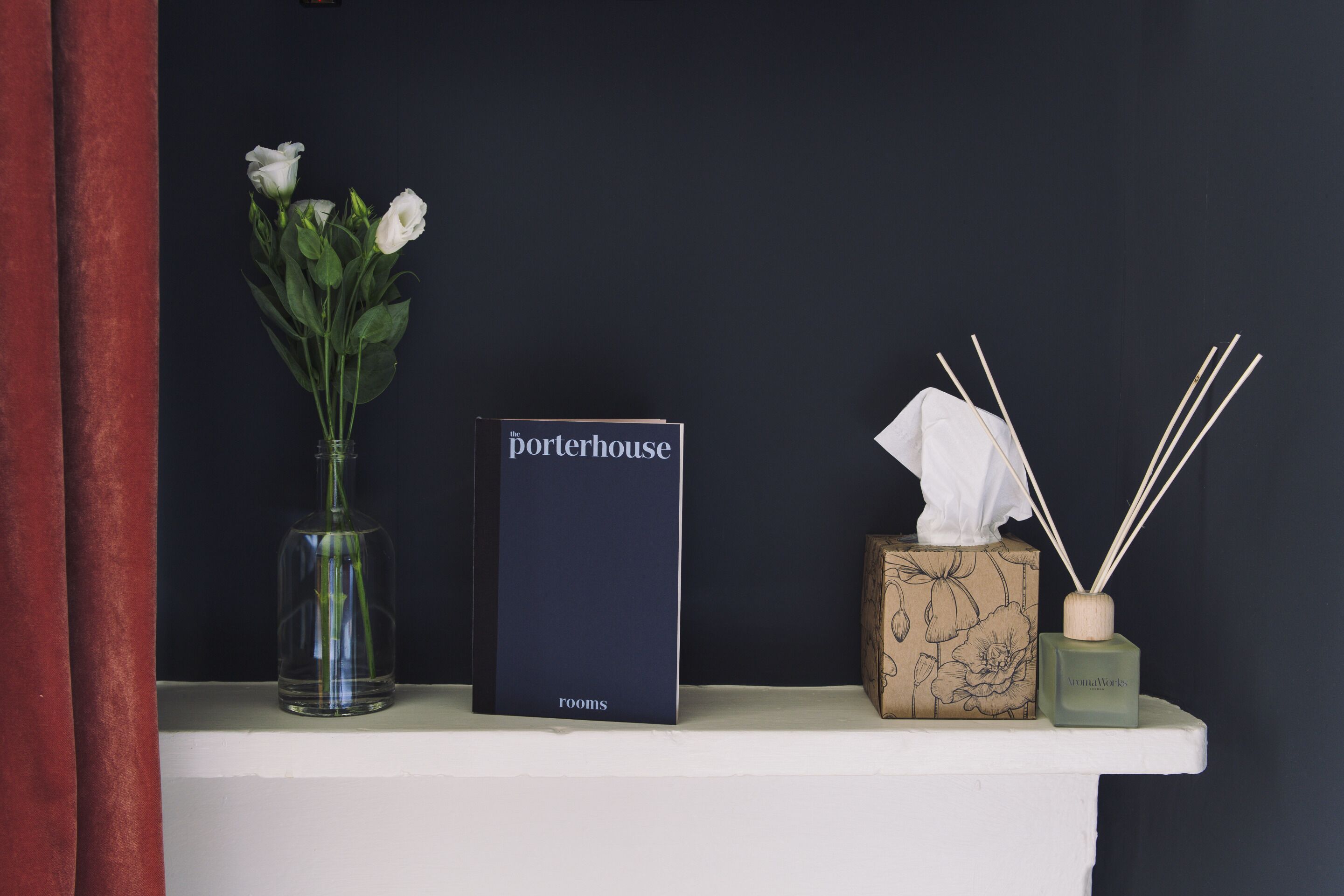 A shelf with a vase of white flowers, a box labeled 'porterhouse rooms', a tissue box, and reed diffuser sticks.
