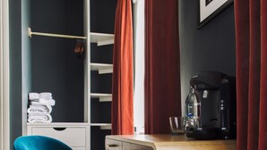 Ein modernes Zimmer mit einem türkisblauen Sessel, einem Holztisch mit Kaffeemaschine und einem Schrank mit roten Vorhängen.