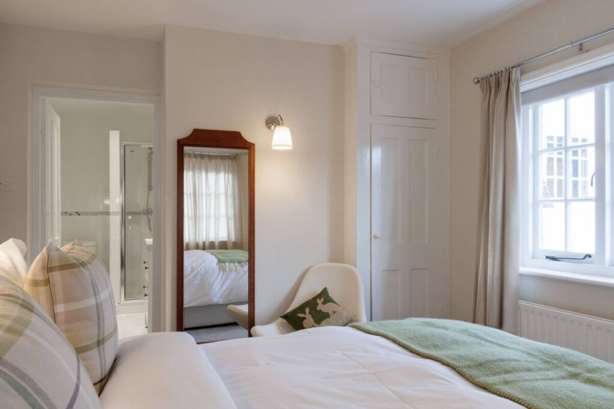 Quarto de hotel com uma cama, uma cadeira, um espelho, uma janela com cortinas e uma porta que dá para um banheiro.