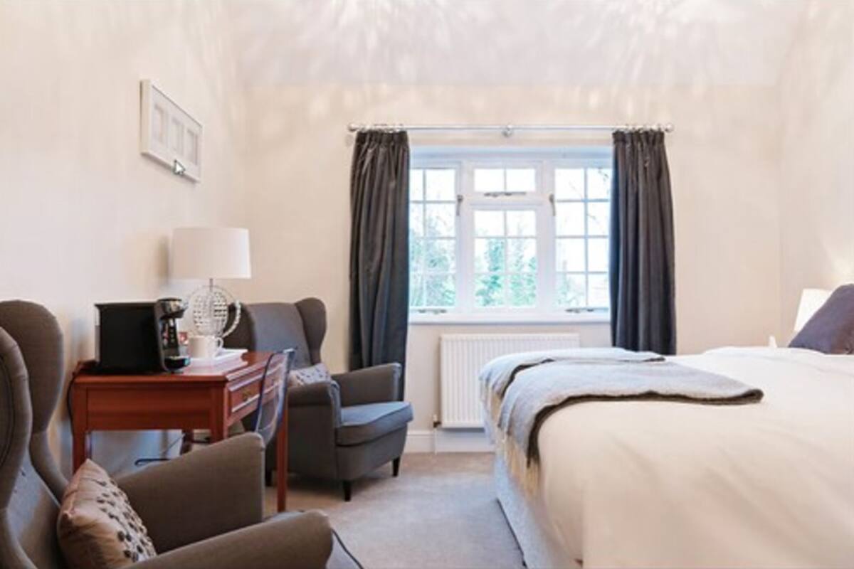 Quarto de hotel com cama, escrivaninha com luminária, cadeira e janela com cortinas.