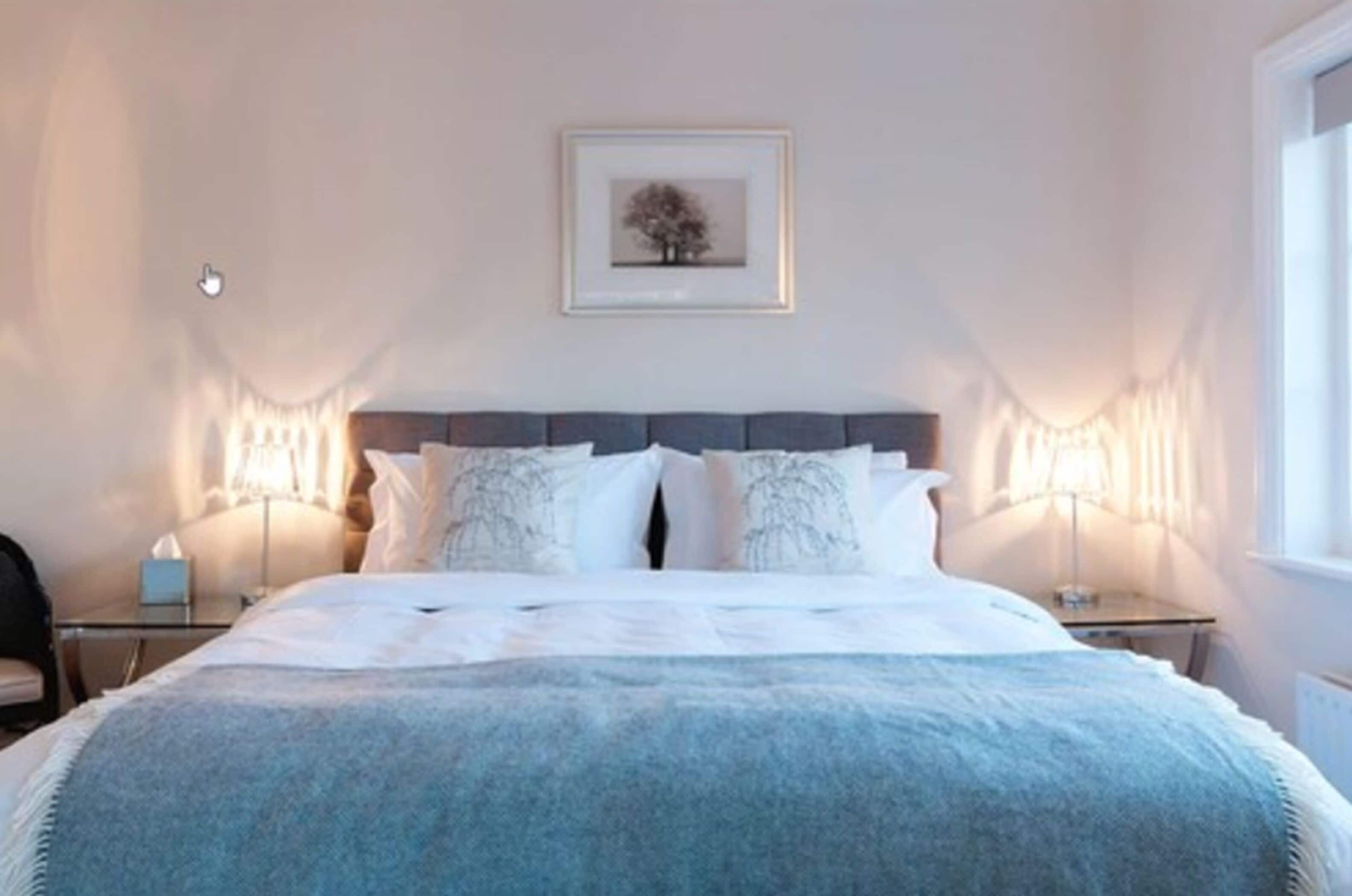 Ein Schlafzimmer mit einem großen Bett, zwei Nachttischen, einem Beistelltisch, einem gerahmten Bild an der Wand und einem Fenster mit Vorhängen.