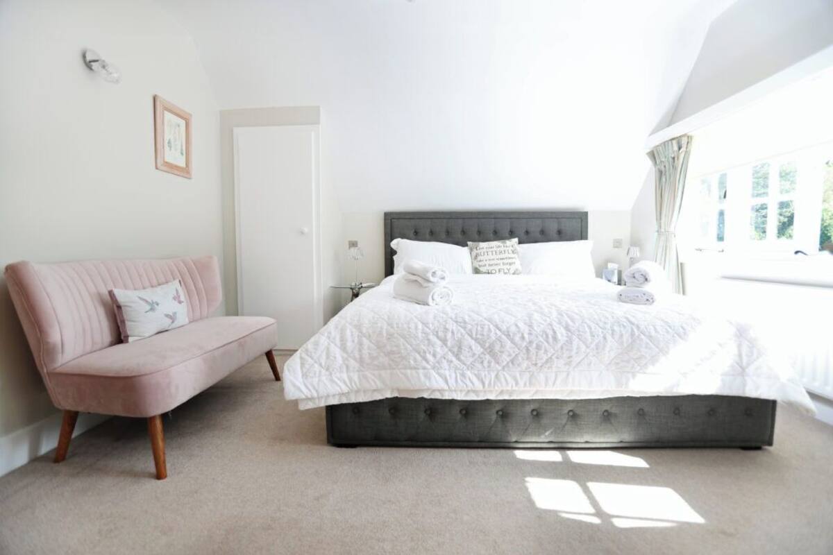 Quarto com cama, sofá rosa e janela com cortinas.