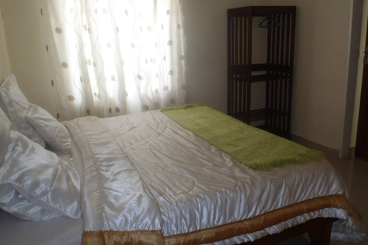 A neatly made bed with a white duvet and a green throw blanket, a wooden wardrobe, and a window with a curtain.