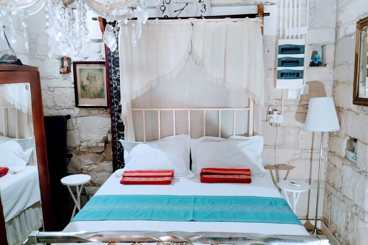 A bed with white linens and red and blue pillows, a small bedside table, a lamp, and a wall clock.