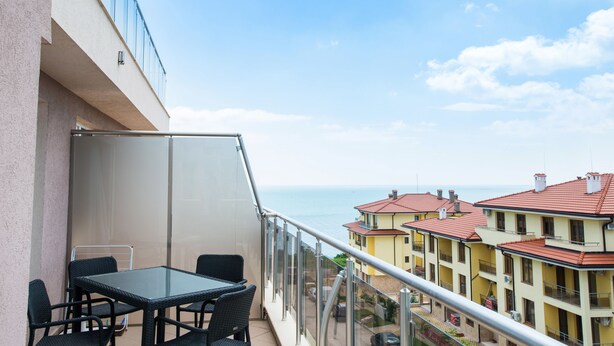 Un balcon avec une table et des chaises en verre, donnant sur une zone côtière avec des bâtiments.