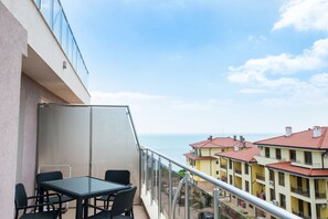 Uma varanda com mesa e cadeiras de vidro, com vista para uma área costeira com edifícios.