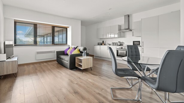 A modern living room with a sofa, a glass coffee table, and a TV. There is a kitchen area with cabinets and appliances. - Hamilton Grace - The Quadrant (Swindon)