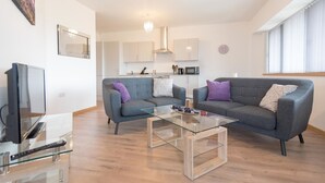 A modern living room with two grey sofas, a glass coffee table, a flat-screen TV, and a kitchen in the background. - Hamilton Grace - The Quadrant (Swindon)